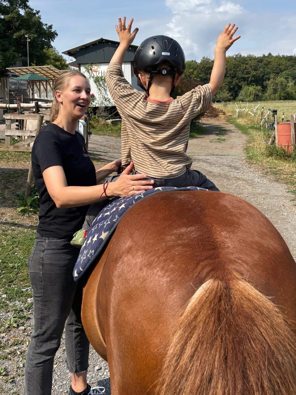 Kind streckt auf dem Pony beide Arme in die Luft, eine Mitarbeiterin lacht und stützt es