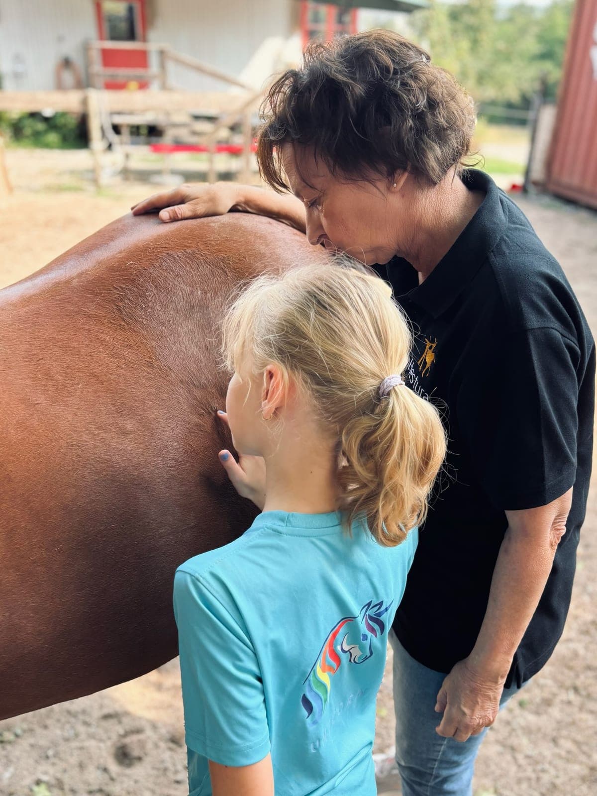 Ein Kind lehnt sich an ein Pferd, eine Mitarbeiterin legt ruhig die Hand auf den Pferderücken